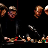 Sō Percussion, four musicians dressed in all black, are gathered around an instrument and playing it with colorful mallets.