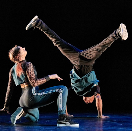 Two Ephrat Asherie dancers are in break dance movements. One is squatted on one knee facing the sky, and the other lifts themselves from the floor with one arm.