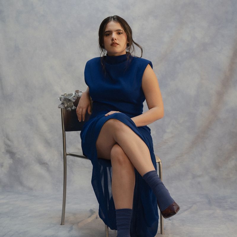 A woman with dark hair in a blue dress sits cross legged in front of a grey backdrop.