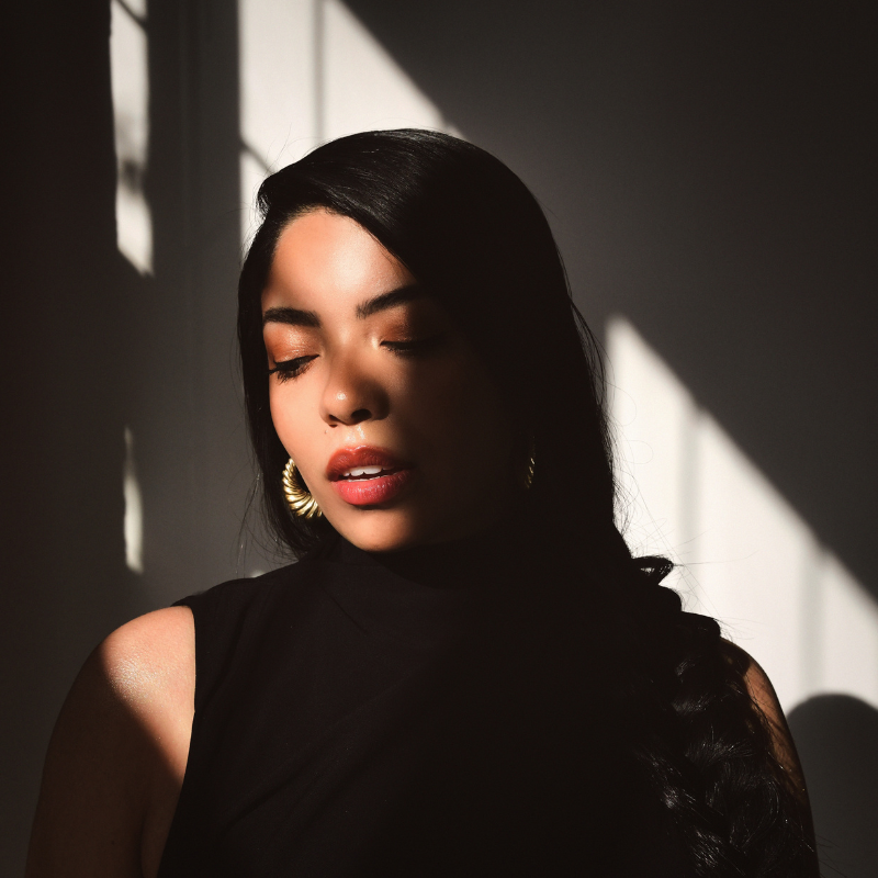 A woman stands in a black dress with light spilling in from the window.